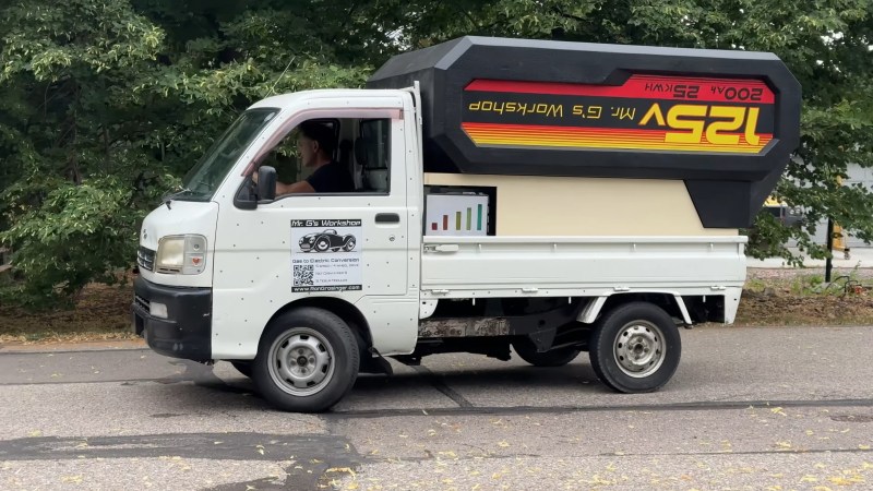 A small white work truck sitting on a faded road with trees in the background. In its bed is what looks like an enormous drill battery in an upside down position. The battery is black with red and yellow stripes. It has the words 125V, 500 Ah, 52 kWh and Mr. G's Workshop emblazoned on the side.