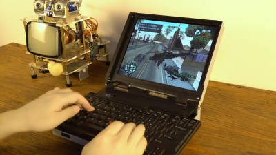 A pair of hands play GTA on a butterfly keyboard-having laptop as a robot looks on.