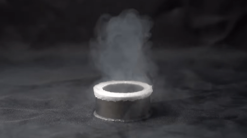 A cylindrical neodymium-iron-boron magnet in shown on a grey background. A white line of boiling liquid surrounds the top edge of the magnet, and a cloud of condensing vapor hangs over it.
