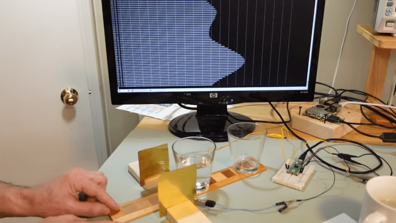 A man's hand is holding a thin wooden board, which runs between two vertical metal plates. On the board are two glasses, one filled with water. A computer screen is behind all this, showing a series of white bars. The series of bars shows two peaks.