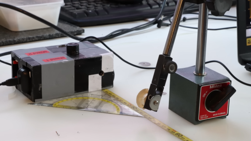 A lead box with a small aperture sits on a desk. A ruler leads away from it. A small disk on a stand is held in front of the aperture.