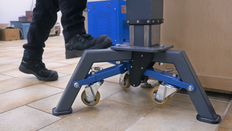A booted foot is seen descending on a foot pedal attached to a lever underneath a three-legged stand. Three levers extend from the legs, each with a wheel on it, and attach to the central foot plate.