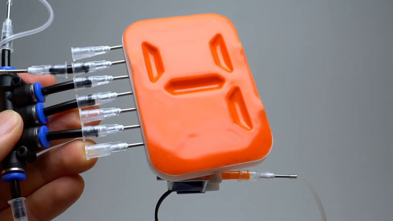 An orange silicone sheet is shown in front, with depressions in the shape of a 7-segment character "4". A man's hand is holding a pipe leading to a series of needles, which enter the block behind the silicone sheet.