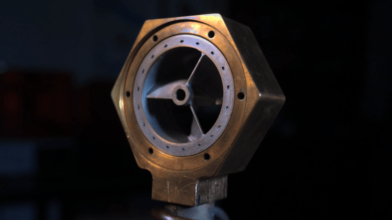 A hexagonal brass enclosure surrounds an aluminium fan with three blades. The fan has an integrated outer rim with a series of small holes around the rim.