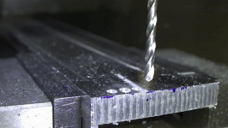 A drill bit, not apparently rotating, descends toward a block of aluminium and throws up aluminium shavings as it passes through the block.