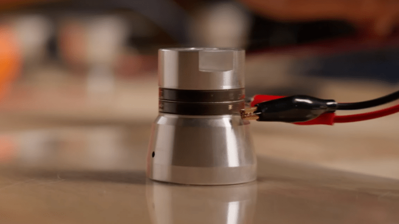 An ultrasonic transducer with two wires attached to it by alligator clips floats very slightly suspended over a glass surface.