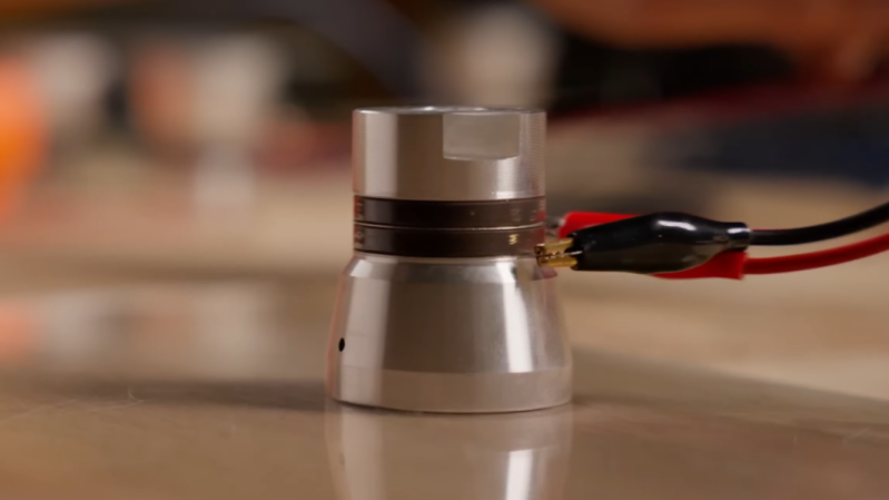 An ultrasonic transducer with two wires attached to it by alligator clips floats very slightly suspended over a glass surface.