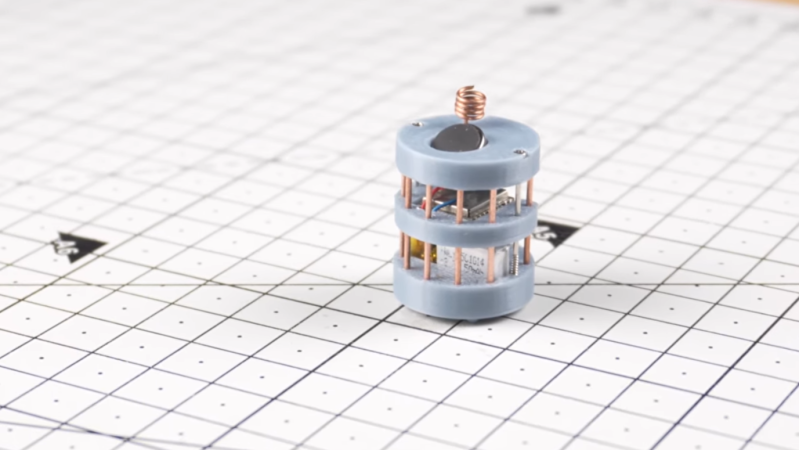 A cylindrical grey robot sits on a white mat. The robot is made of three grey sections connected by a series of radially-arranged copper pillars.