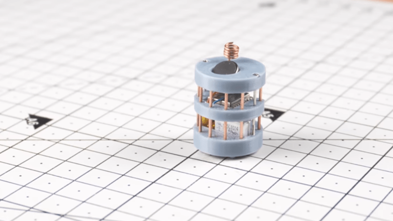 A cylindrical grey robot sits on a white mat. The robot is made of three grey sections connected by a series of radially-arranged copper pillars.