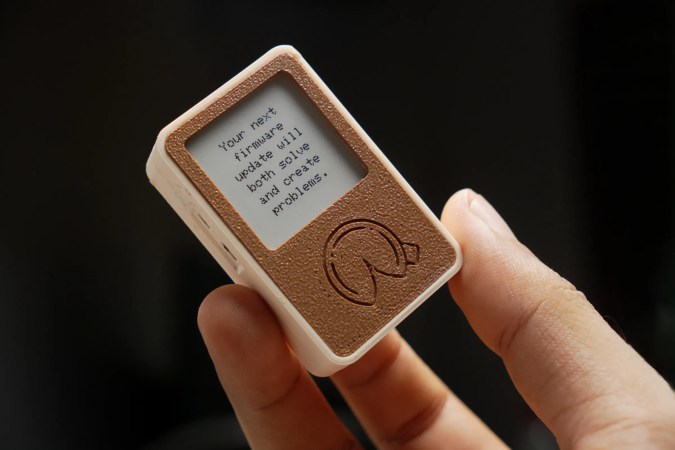 A tiny, rectangular, 3D-printed box with an e-paper display and a fortune cookie design beneath it. The fortune reads: "Your next firmware update will both solve and create problems."
