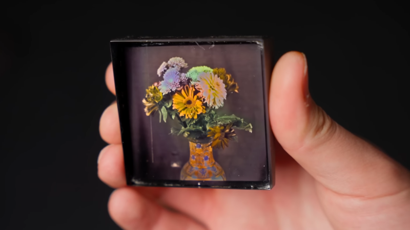 A man's hand is shown holding a color photograph of a vase of flowers against a black background.