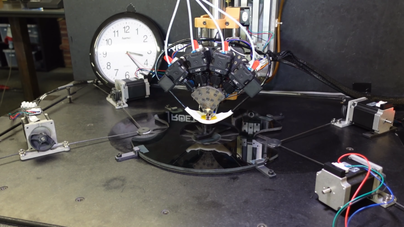A 3D printer hotend with four filament leads in positioned on an arm above a hole in a glass plate. Wires lead from a carbon fiber frame under the glass to four stepper motors with pulleys.