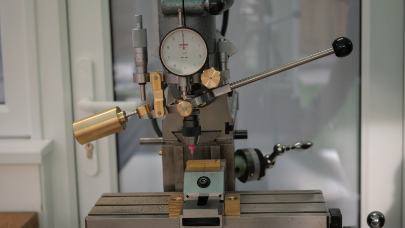 A mechanism made of brass and steel is attached to a milling machine. It has a long lever extending from the right side, a counterweight attached to the left side, and an indicator gauge in the middle.