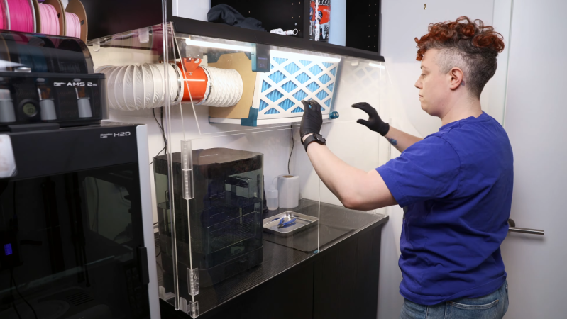 A person is standing in front of an acrylic enclosure, lowering a door on the enclosure. The enclosure contains the space between two sets of cabinets, and has three doors on the front. Inside the enclosure is an air filter and a washing station.
