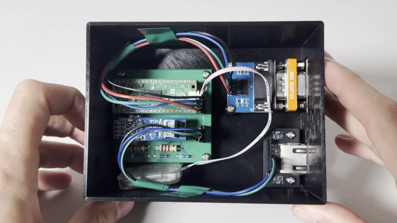 A black plastic box is shown, with a green circuit board inside. The circuit board is wired to an RS-232 connector and an RJ-45 connector.