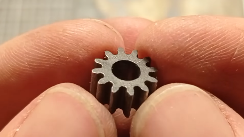 A fine steel gear is shown held between a man's fingertips.