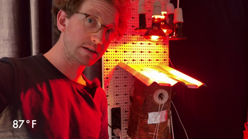 A white man in a dark t-shirt and glasses stands next to a pegboard. On the pegboard is a cylindrical wooden bird house with a small piece of metal roof attached to the top. A set of heat lamps are suspended above and give the image a reddish-orange glow. 87˚F is in white text in the lower left corner.