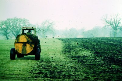 The rear view of a tractor towing a manure spreader driving away from the viewer while spreading manure onto a grass field. It's a misty winter day, and leafless trees are visible in the distance.