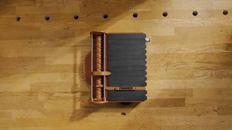 A device with a brown plastic gear cylinder and indicator on the left and a series of black pieces approximately the size of tongue depressors sits on a butcher block workbench. A thin brown divider with the word "Finished" sits between sets of the presumably chalkboard tongue depressors.