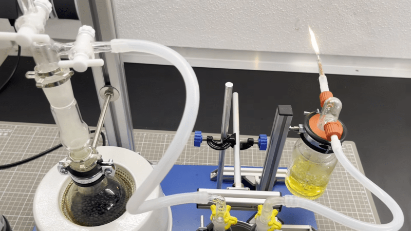 A boiling flask is mounted in a heating manted, with a tube leading from it to a U-shaped tube. From here, the tube continues to a bottle of yellow fluid, from which another tube emerges. A flame is emitted from this last tube.