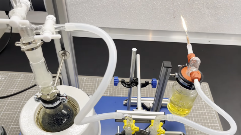 A boiling flask is mounted in a heating manted, with a tube leading from it to a U-shaped tube. From here, the tube continues to a bottle of yellow fluid, from which another tube emerges. A flame is emitted from this last tube.