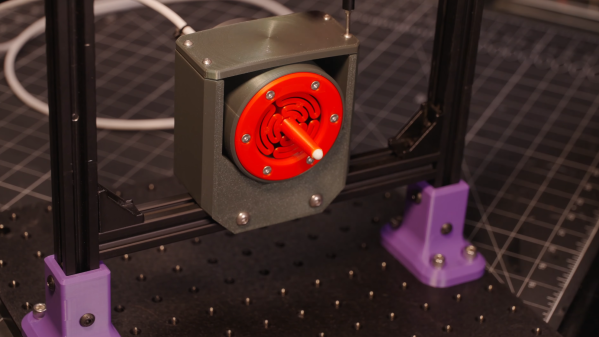 A grey box surrounding a circular red component is mounted on an aluminium extrusion frame. The circular red face has a protrusion extending from it with a white ball bearing at the tip.