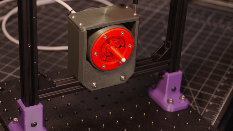 A grey box surrounding a circular red component is mounted on an aluminium extrusion frame. The circular red face has a protrusion extending from it with a white ball bearing at the tip.