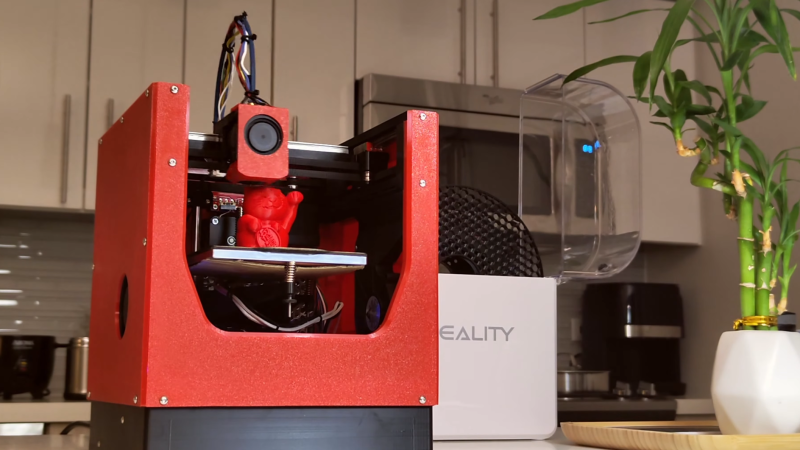 A small, orange 3D printer is shown on a desk with a filament dry box. The printer is printing a waving cat figurine. The printer is a CoreXY configuration, and the side panels are 3D-printed orange plastic.