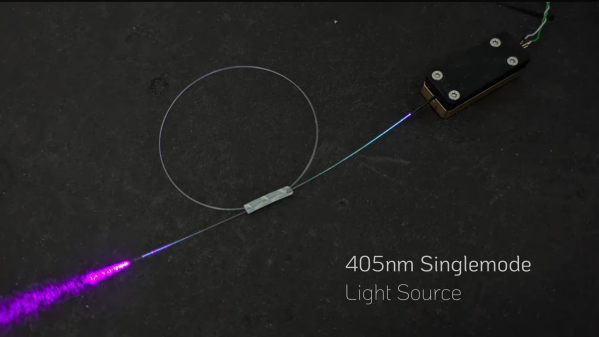 A rectangular black box is shown, connected to a coil of fiber-optic wire. Out of the end of the fiber, purple light is emitted. A label in the lower right corner says "405nm Singlemode Light Source".