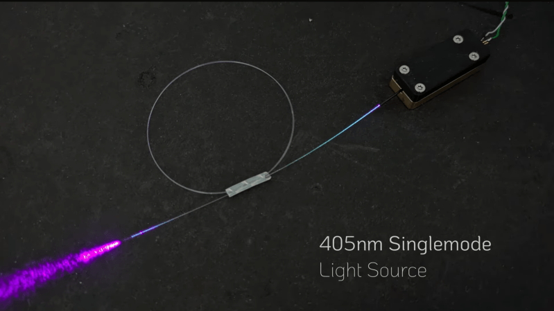 A rectangular black box is shown, connected to a coil of fiber-optic wire. Out of the end of the fiber, purple light is emitted. A label in the lower right corner says "405nm Singlemode Light Source".