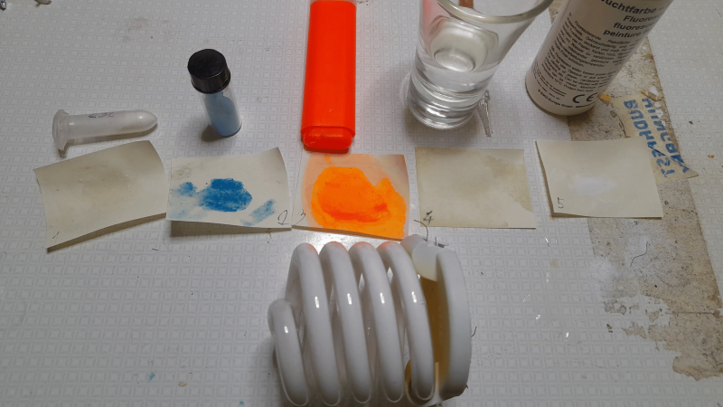 A selection of materials sits on a counter. There is a fluorescent light bulb, two papers stained with dyes, and a few other pieces of paper with no obvious staining.