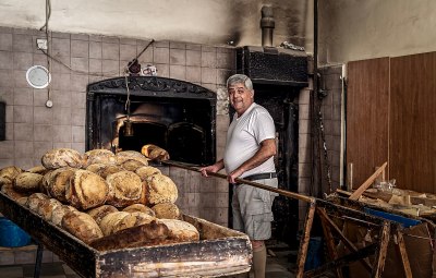 A baker stands next to the opening of his oven, a fresh loaf of bread on his paddle. In the foreground is a stack of loaves.