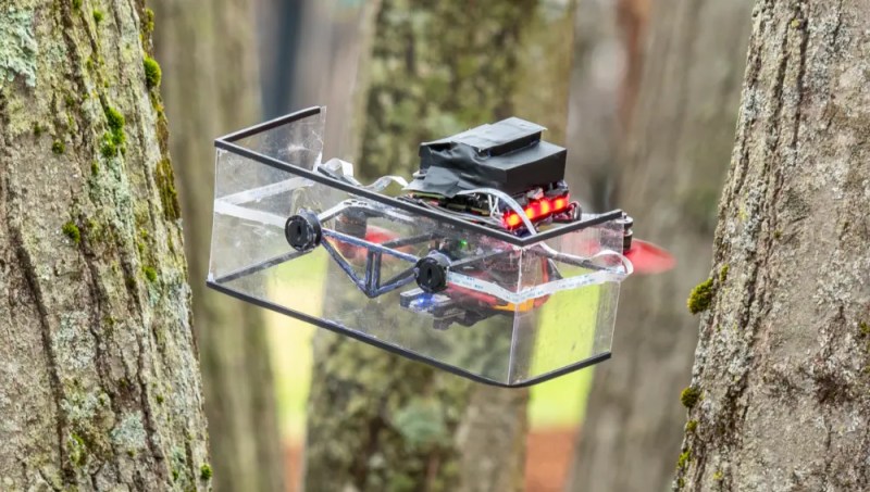 A quadcopter with a clear, U-shaped shield up front and red propellers navigates between a close stand of moss-covered trees.