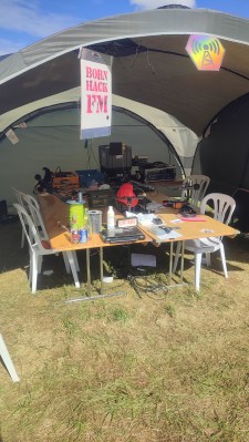 A view of a tent shelter in bright sunlight with studio equipment visible on a table in it. There's a sign: "BornHack FM".