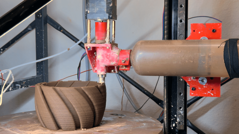 A clay vase sits in the center of a circular table, with an extruder in contact with the top surface. The extruder has a tube containing clay on the right side, with a motor mounted above an auger over the main nozzle.