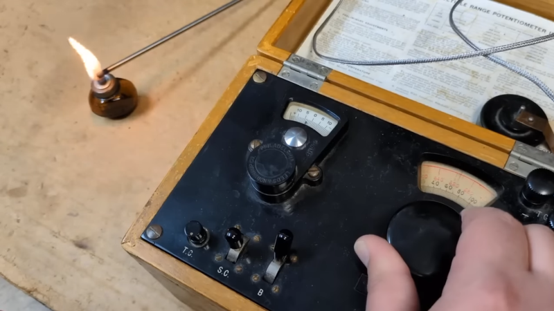 A man's hand is shown adjusting a black Bakelite dial on the front panel of an instrument. The instrument is contained in a wooden box, and to the left of the box, a thermocouple is inserted into the flame of an alcohol burner.