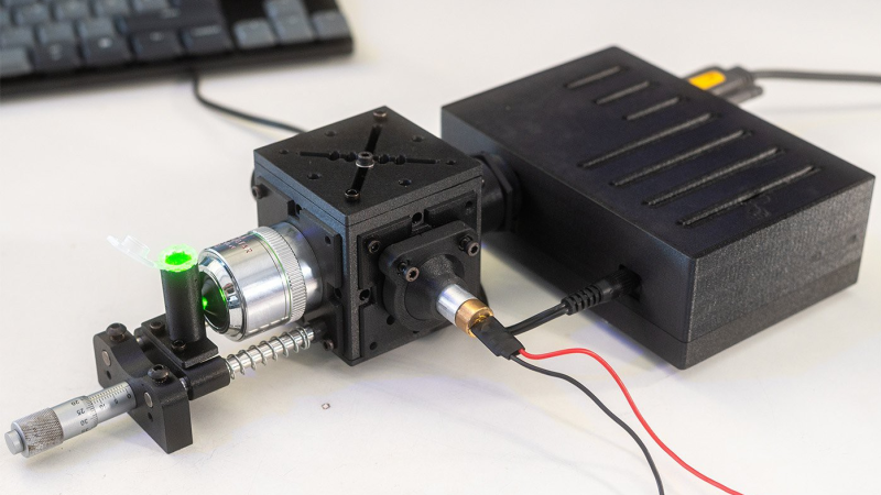 A black plastic cube is shown in front of another, larger rectangular black plastic box. The plastic cube has a silver microscope objective protruding from one side, with green light being emitted from it into a small plastic tube held on a positioning stage.