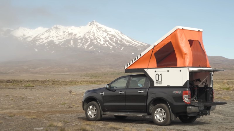A black pickup sits in front of a snow-capped mountain range in the background. It has a white camper shell with a sci-fi looking "01" emblazoned on the side in black text. An orange, wedge-shaped tent pops up above the campershell and truck roof.