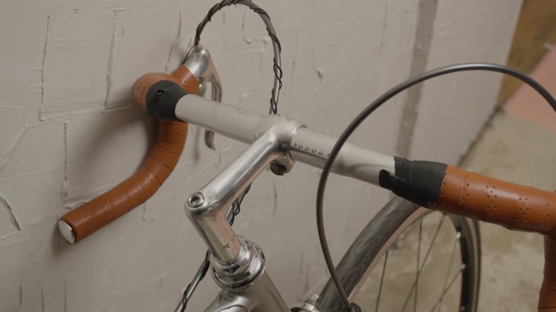 Brown wrapped chrome handlebars leaning against a light grey wall. Near the stem, a series of four small pins protrude from the bars to indicate a full battery charge of 100%.
