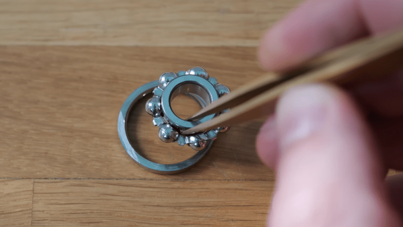 A man's hand is shown holding brass-colored tweezers. In the tweezers are held the inner race and the ball cage of a ball bearing, with half of the outer race lying below them on a table.