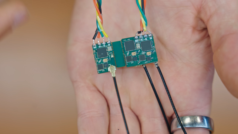 A man's hand is shown holding two small circuit boards, joined together by a central bridge. Each circuit board is connected to four wires and a coaxial antenna cable.