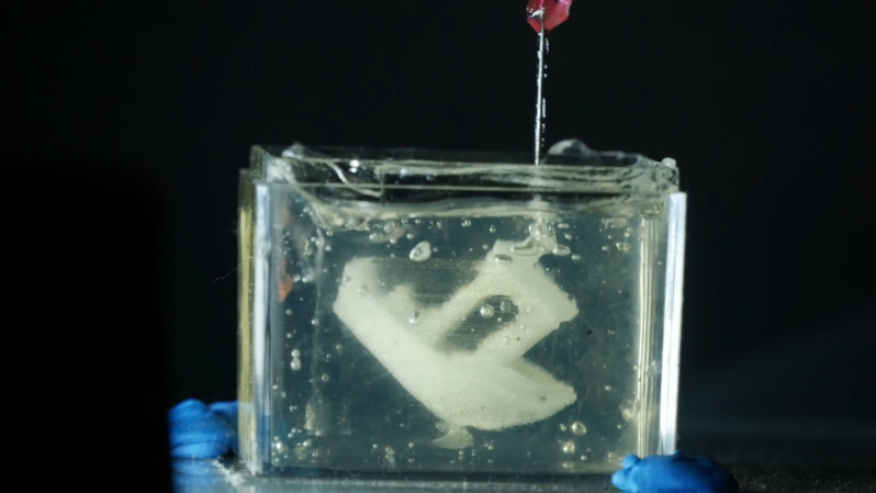 A clear tank of gel is shown against a black background. In the gel, the shape of a white plastic boat is suspended. A coarse-tipped needle dips into the top of the gel bath.