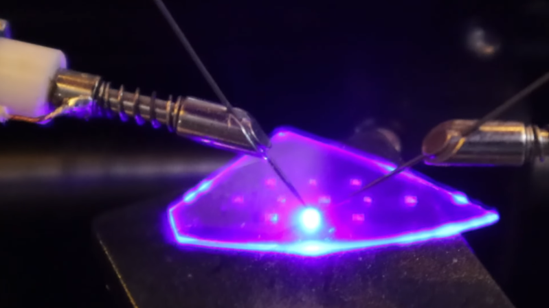 An irregular shard of a crystal wafer is set on a table, with two wire probes contacting it. Between the probes, a bright blue-purple light glows, illuminating the rest of the wafer.