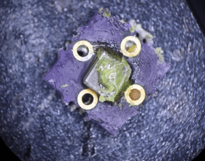 A square, purple PCB is shown under a microscope. The PCB has four vias surrounding a transparent chip, which has a blob of translucent yellow material on top of it.