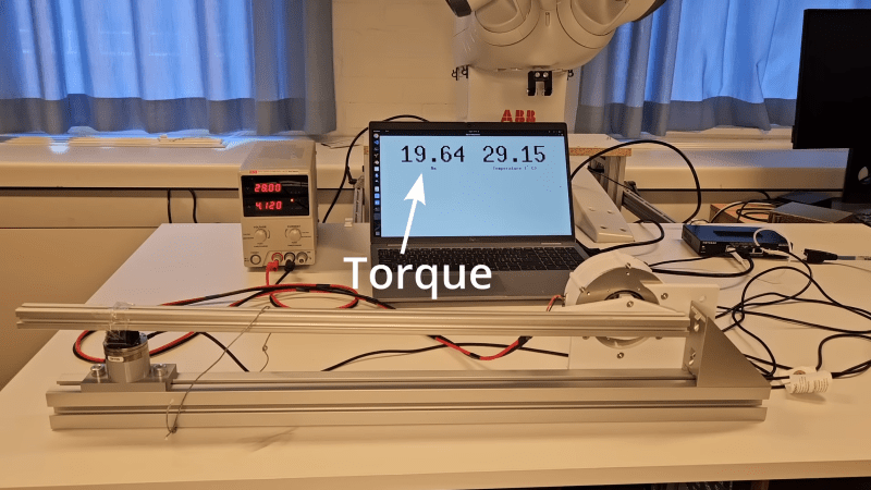 A test setup for an actuator is shown on a benchtop. A power supply connects to a cylindrical actuator, which has an arm pressing against a load cell. The load cell's output is shown on a computer screen, which is labelled "Torque".