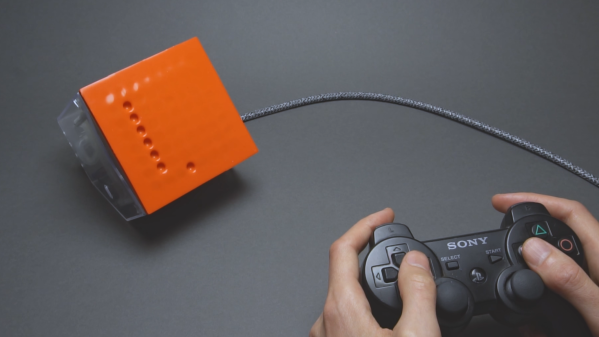 A man’s hands are shown holding a video game controller. A cable runs to a box with an orange front surface, which has a series of divots arranged in a points on a grid. These divots form a vertical line, with one other divot to the right of and below the line.