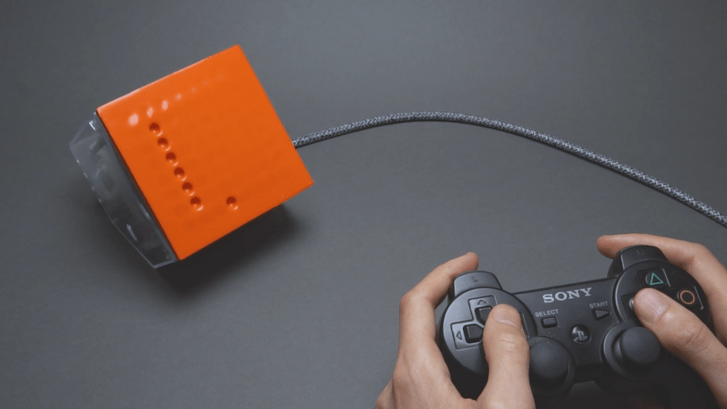 A man’s hands are shown holding a video game controller. A cable runs to a box with an orange front surface, which has a series of divots arranged in a points on a grid. These divots form a vertical line, with one other divot to the right of and below the line.