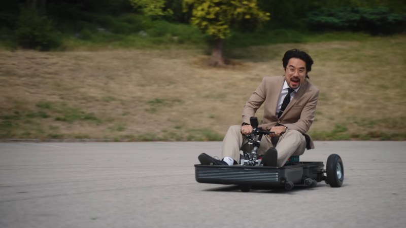 A man in a beige, vintage 90s suit rides a very small go cart with a look of terror and/or elation on his face.