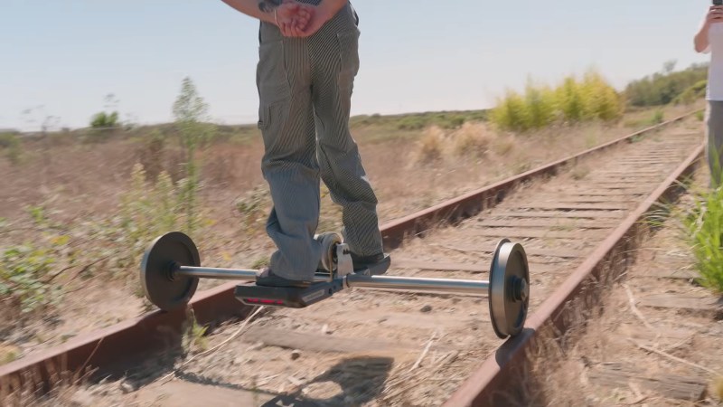 A man in white and black railroad overalls rides a short one wheel platform suspended between two rail wheels on a steel axle.