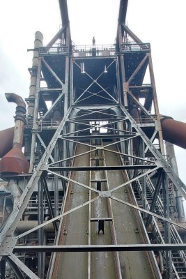 A ramp covered in a latice of girders slopes steeply upwards. Visible on it are some rails, and at the top is the top of the furnace.